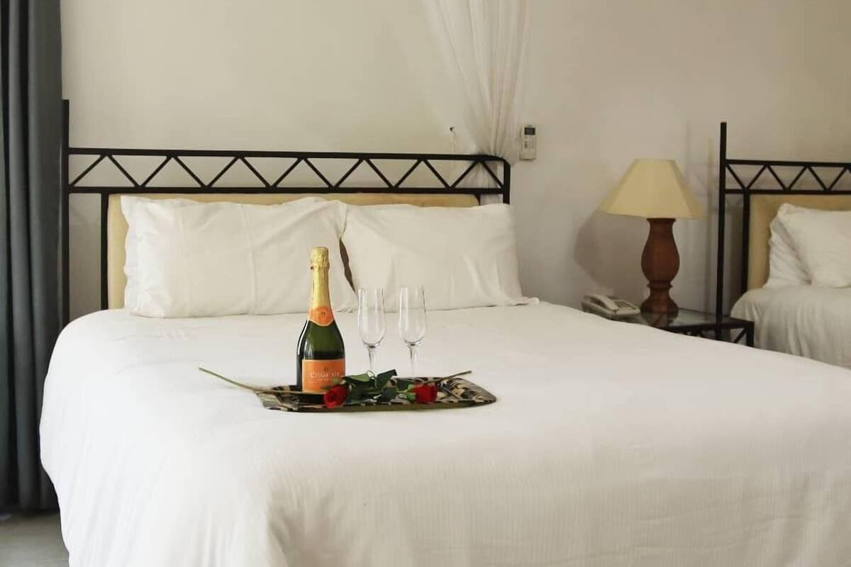 Una habitación de hotel con una cama, una mesita de noche con una lámpara y una botella de champán sobre la cama.