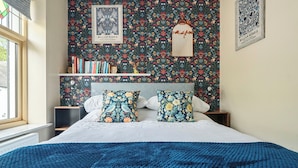 A bedroom with a floral wallpaper, a bookshelf, a bed with a blue blanket, and a window.