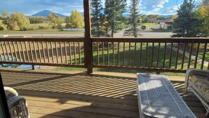 A wooden deck with a covered area, two wicker chairs, and a view of a road and trees. - Grandote Lodge (La Veta)