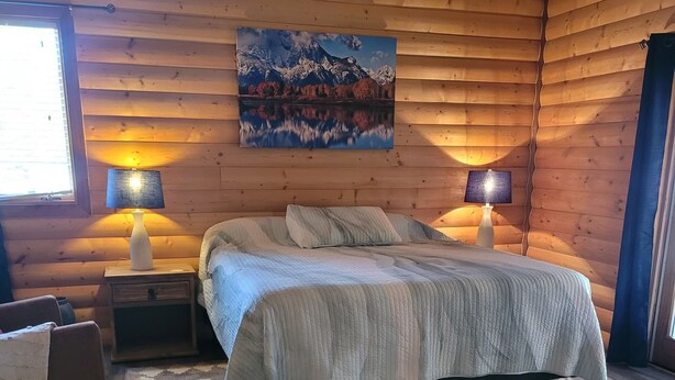 A cozy log cabin bedroom with a bed, two bedside lamps, a rug, and a painting of a mountain landscape. - Grandote Lodge (La Veta)