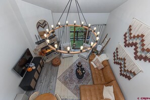 A modern living room with a fireplace, a large chandelier, and a view of the outdoors. - Hot Tub, Fishing/Hiking Close, 30 Min to Silverton! (Durango)