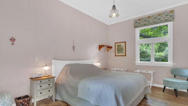 A bedroom with a bed, bedside tables, a lamp, a window with a view of greenery, a chair, and a small rug. - Rose Cottage - Tranquil retreat 
 (Masterton)