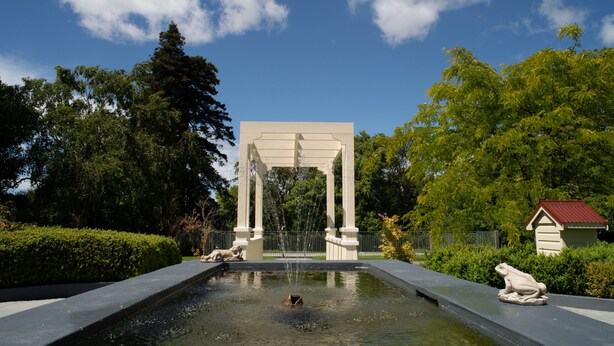 A fountain with a statue of a frog, surrounded by greenery and a white pavilion.