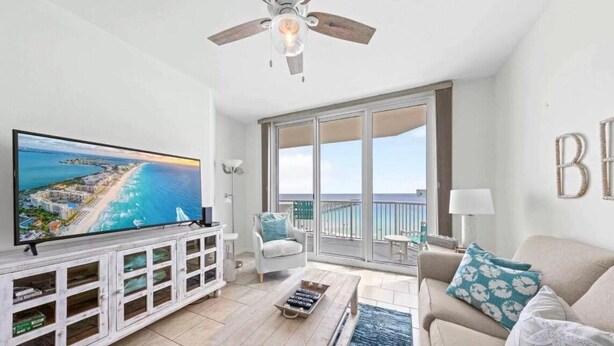 Een ruime woonkamer met een grote televisie, een plafondventilator, een balkon met uitzicht op zee, en een gezellige zithoek.
