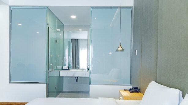 Une chambre d’hôtel moderne équipée d’un lit, d’une table de chevet, d’un téléphone et d’une salle de bain fermée par des parois de verre, avec un miroir et un lavabo.