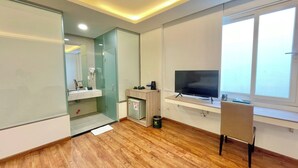 Une chambre d’hôtel moderne avec un plancher en bois, une douche en verre, un lavabo blanc, un petit réfrigérateur, un téléviseur posé sur un meuble et une fenêtre avec vue.