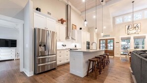 A modern kitchen with stainless steel appliances, a central island with stools, and a dining area with a sofa. - Luxury St. Simons Island Vacation Rental | Private Pool Near Pier Village (Saint Simons Island)