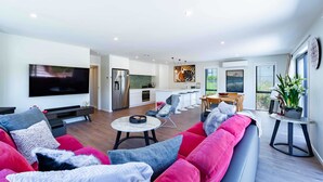 A modern living room with a large flat-screen TV, a red sofa, and a dining area with a kitchen in the background. - Sunset Peaks Wanaka T (Wānaka)