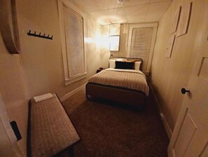 Quarto de hotel estreito com uma cama de solteiro, mesa de cabeceira e janela com persianas.