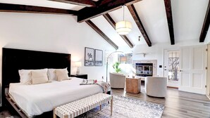 Une chambre avec un grand lit, un banc, un bureau et une chaise. La pièce est dotée d’une porte et d’une fenêtre.