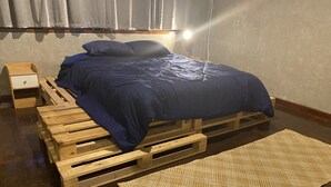 A bed with a blue mattress and wooden frame, a small wooden nightstand, and a woven rug on a wooden floor.