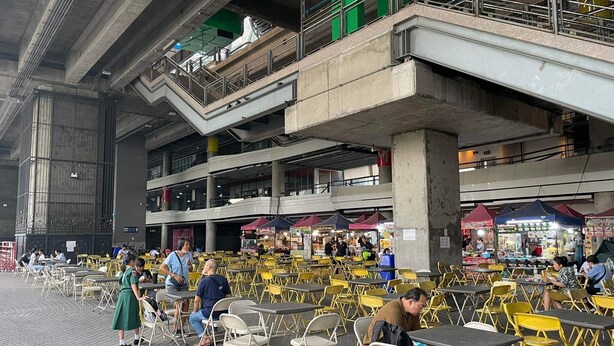 An indoor public space with multiple levels, featuring outdoor seating areas with yellow and white chairs, and various food stalls.