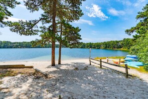A beach with a wooden fence, a row of canoes, and a calm lake surrounded by trees. - Homeport Harwich by the Lakes w/ 3 Kayak's (Harwich)