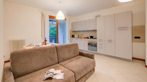 A modern living room with a sofa, a dining table with chairs, a kitchen area with cabinets and appliances, and a large window with curtains. - Magnolia: Laveno Centre 2BR With Terrace (Laveno Mombello)