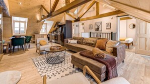 Ein großzügiges Loft mit freiliegenden Holzbalken, einer gemütlichen Sitzecke mit Sofa und Sesseln, einem Esstisch mit Stühlen und einer Küchenzeile.