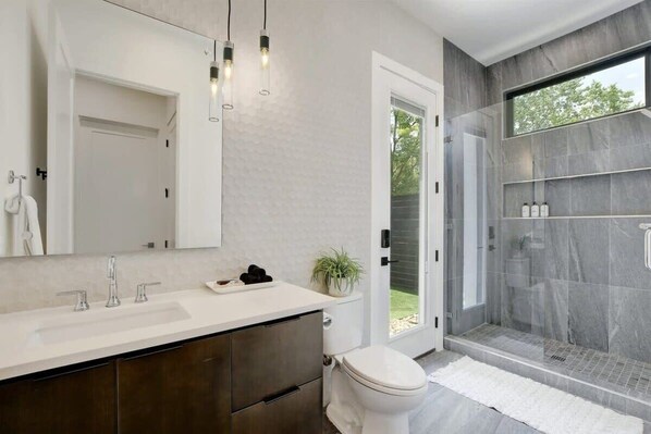 A modern bathroom with a large mirror, a white sink, a toilet, and a glass-enclosed shower area. - 7BR Private Villa with Heated Pool & Outdoor Oasis (Austin)