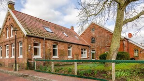 Een klassiek bakstenen gebouw met een schuin dak, een groene omheining en een boom op de voorgrond.