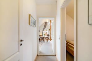 A hallway with a door on the left, a bed on the right, and a dining area in the background.