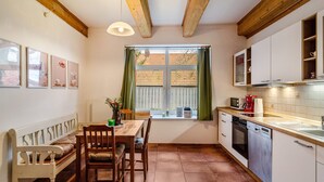 Una cocina con vigas de madera, una mesa de comedor con sillas, una ventana con cortinas y armarios blancos.