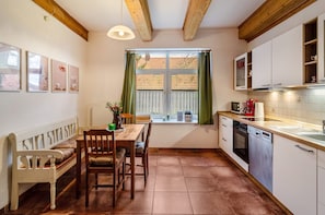 A kitchen with wooden beams, a dining table with chairs, a window with curtains, and white cabinets.