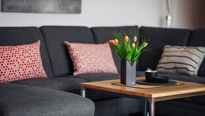 Un salon moderne avec un canapé gris, des coussins à motifs rouges, une table basse en bois et un vase contenant des plantes vertes.