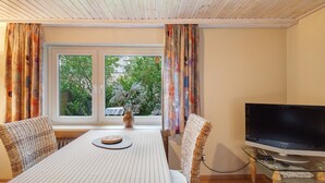 Una habitación con una mesa de comedor, sillas, un televisor sobre una mesa de vidrio y una ventana con cortinas.