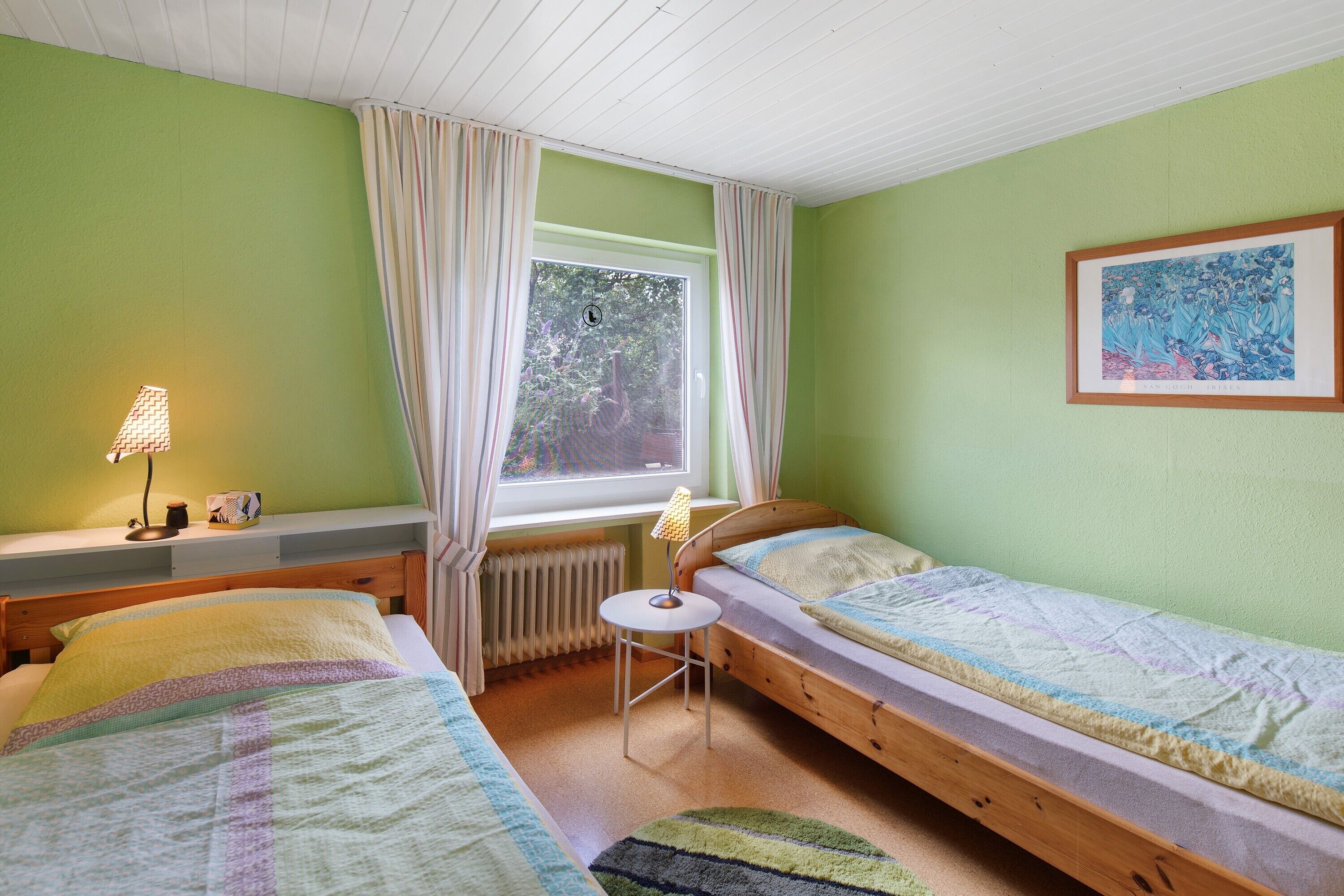 Une chambre avec deux lits, une fenêtre avec des rideaux, un radiateur, une petite table avec une lampe et un tableau encadré au mur.