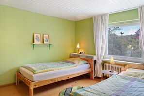 Una habitación con una cama individual, un escritorio pequeño con lámpara, un radiador y una ventana con cortinas.