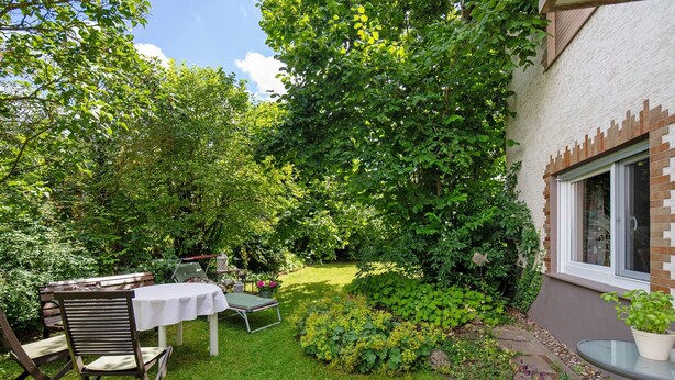 Un jardín con una mesa y sillas, rodeado de abundante follaje y un muro de ladrillo.