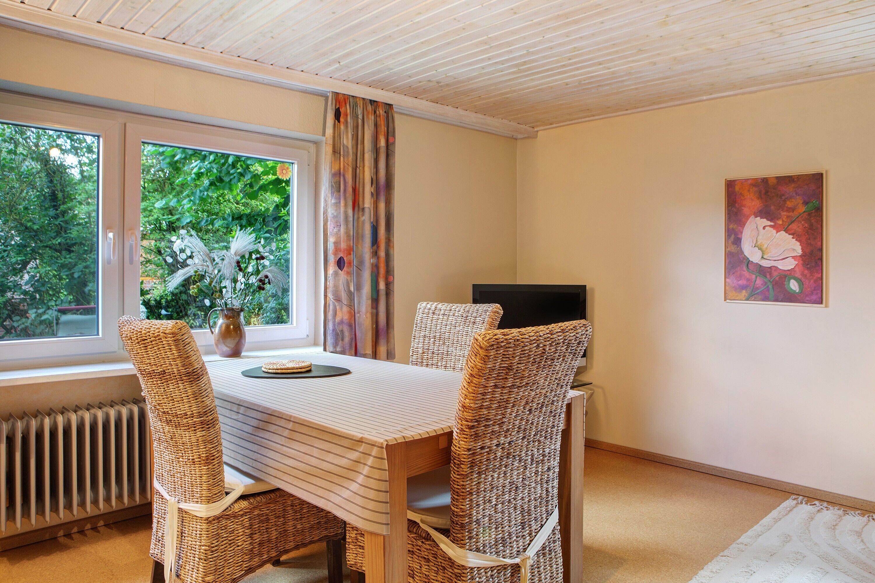 A dining area with a table, wicker chairs, a vase, and a painting on the wall.