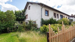 Una casa con techo inclinado, cerca de madera y un jardín con diversas plantas y una silla.