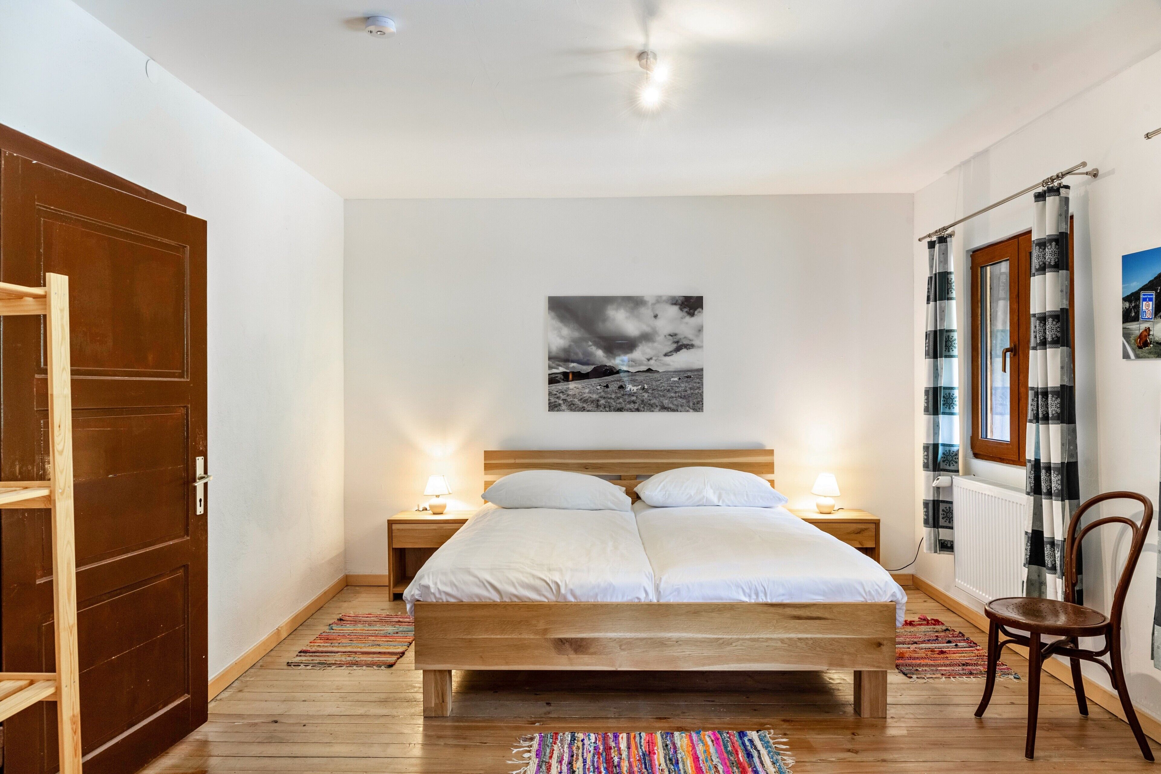 Une chambre avec un lit en bois, deux tables de chevet avec des lampes, un tableau en noir et blanc accroché au mur et une fenêtre avec des rideaux.