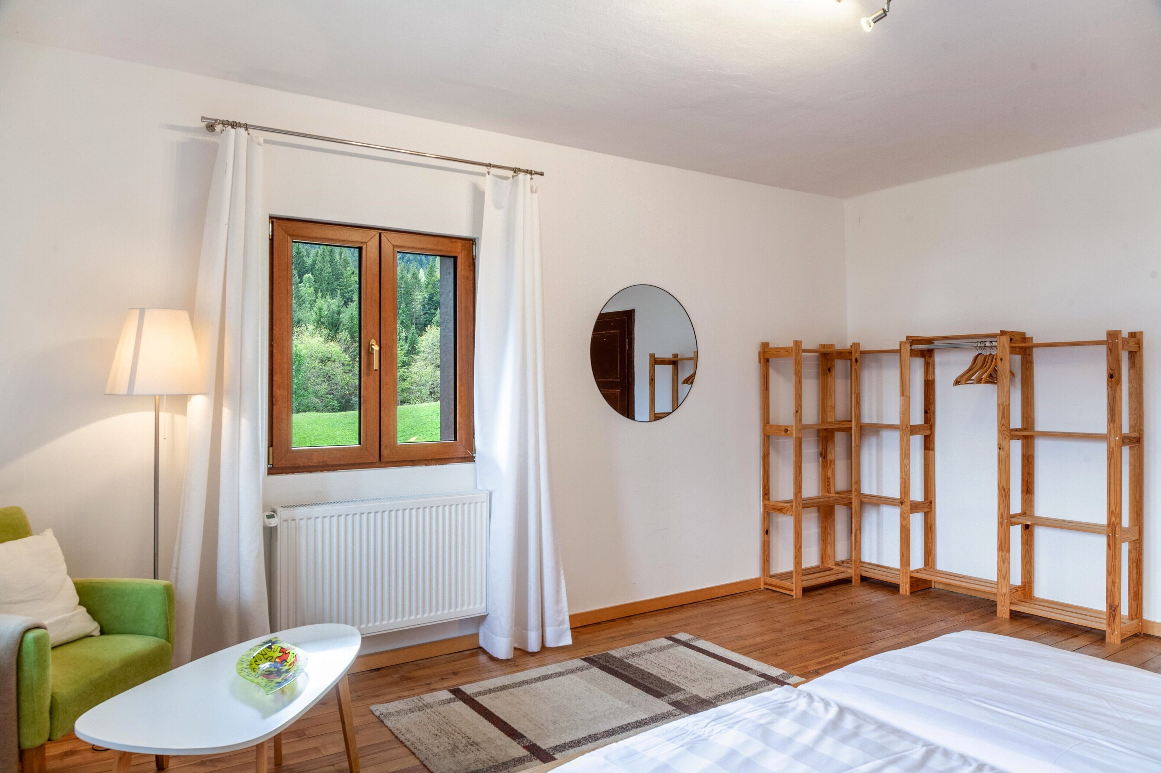 Un dormitorio con una cama, una ventana con cortinas, un radiador, un espejo, una silla verde y una pequeña mesa.