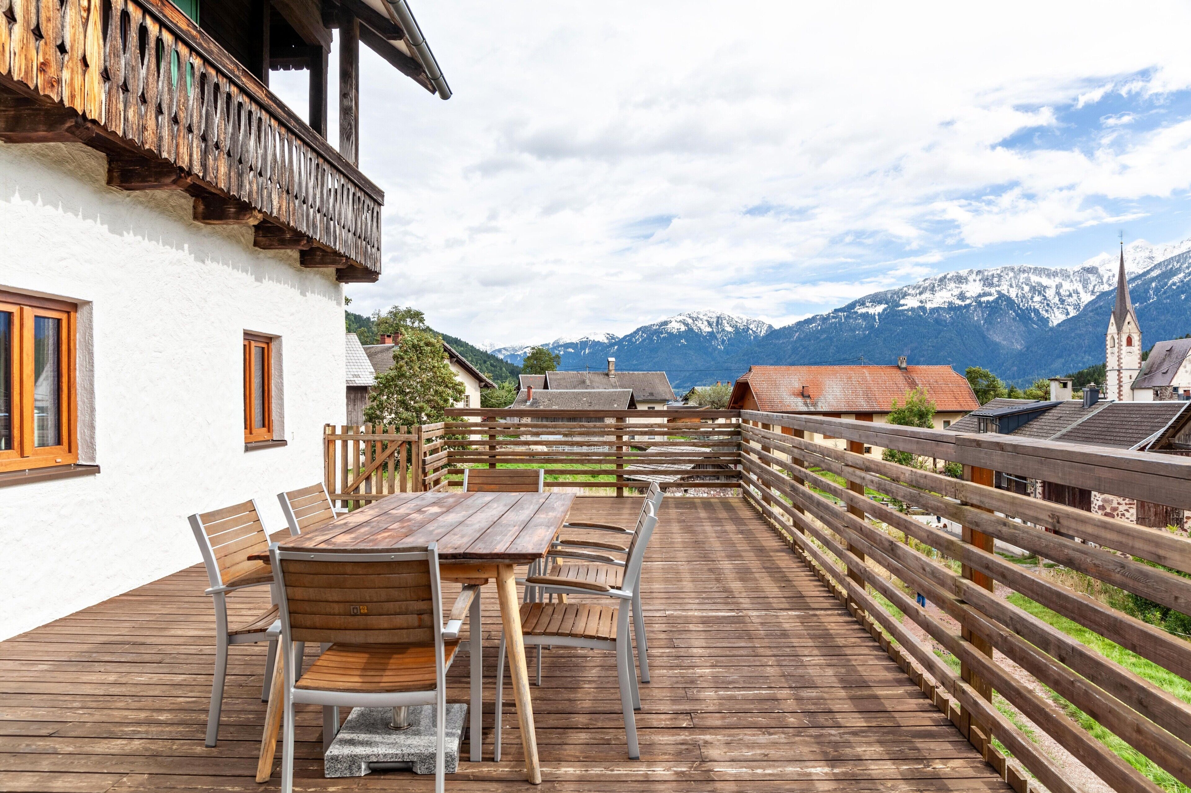 Un patio in legno con tavolo e sedie, un edificio bianco con ringhiere in legno al balcone e una vista sulle montagne sullo sfondo.