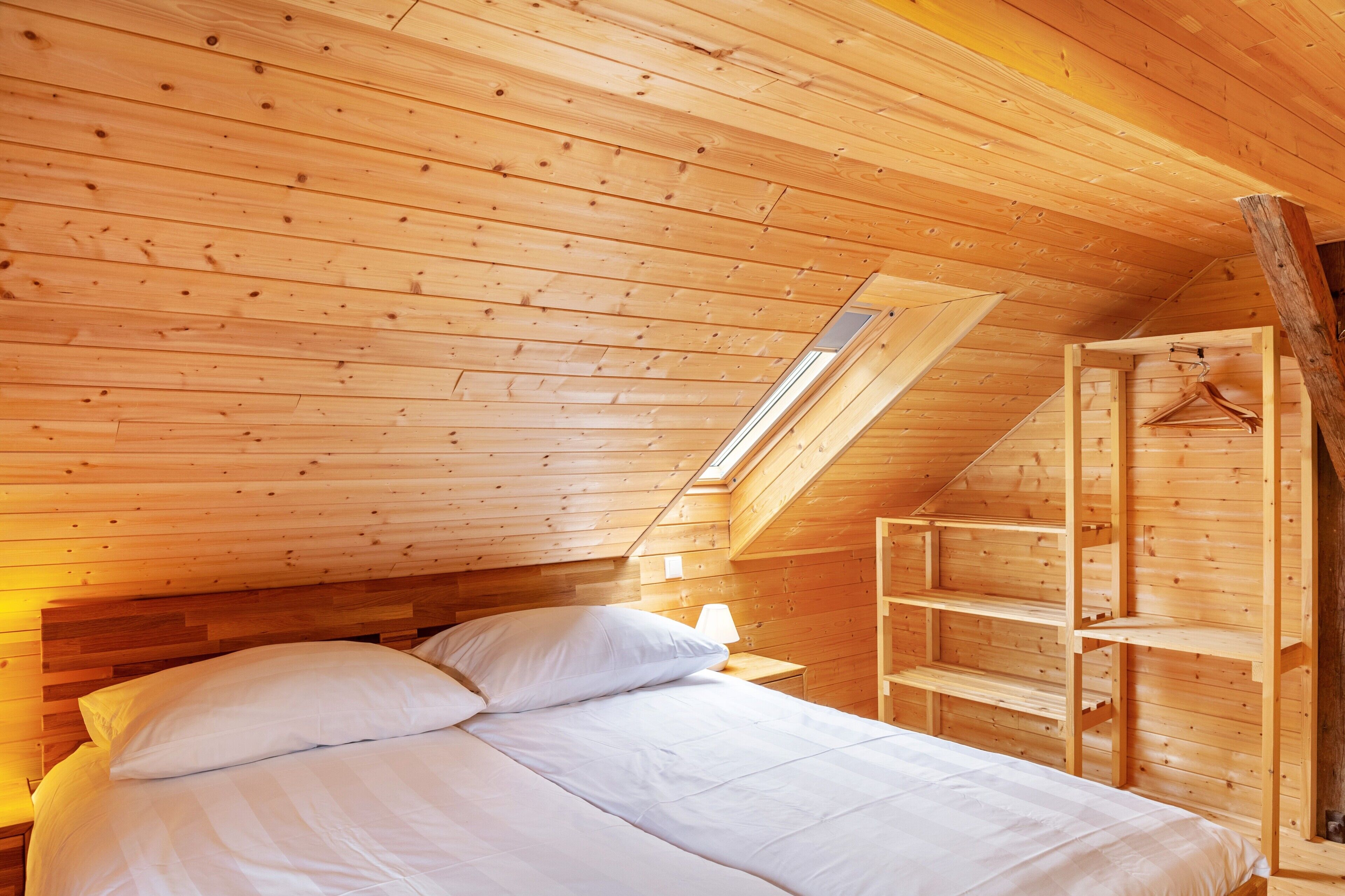 Um quarto com painéis de madeira, uma cama, uma mesinha de cabeceira, um abajur e um claraboia.