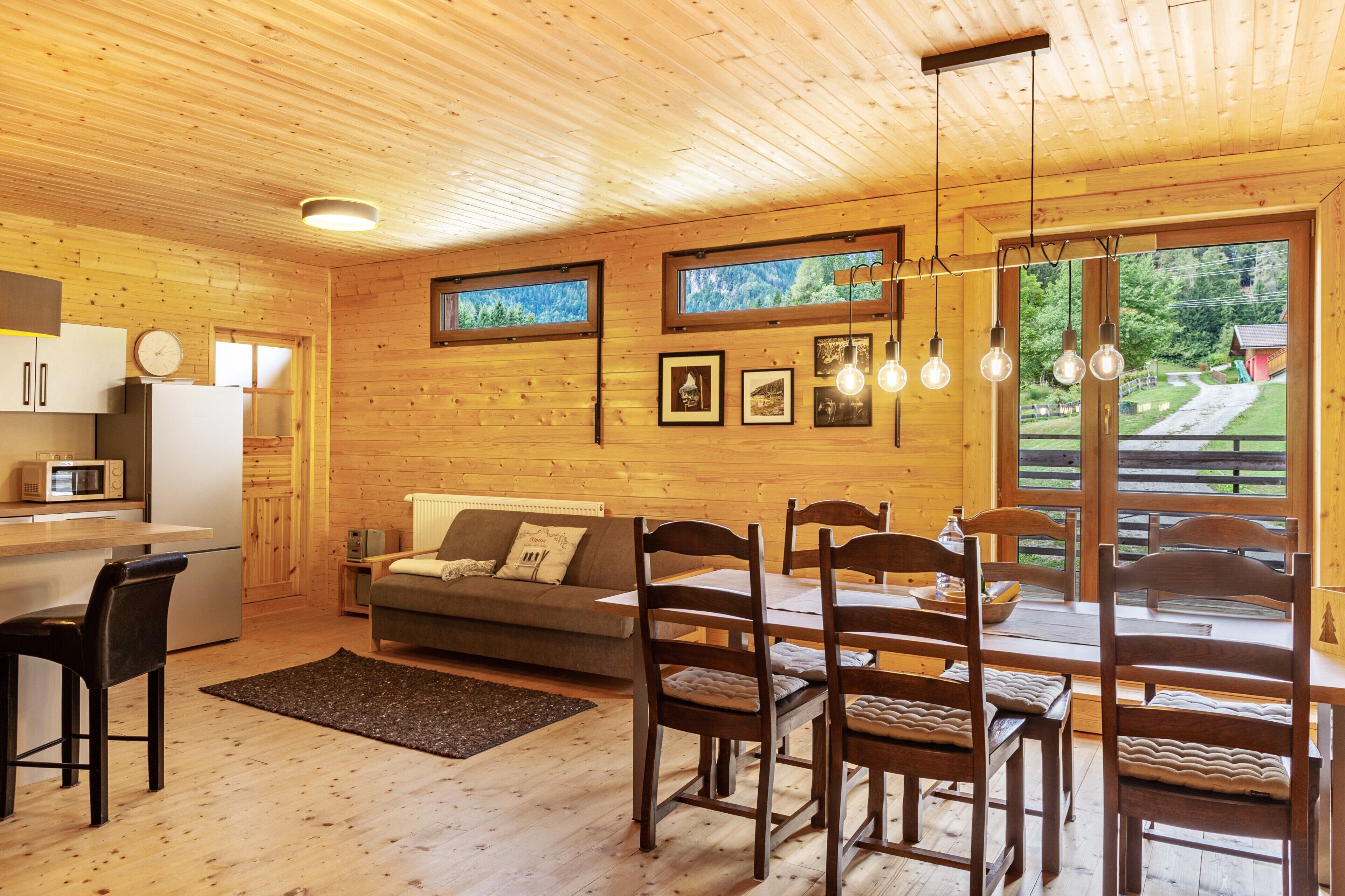A wooden interior with a dining table, chairs, a sofa, a kitchen area, and framed pictures on the wall.