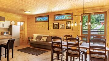 A wooden interior with a dining table, chairs, a sofa, a kitchen area, and framed pictures on the wall.