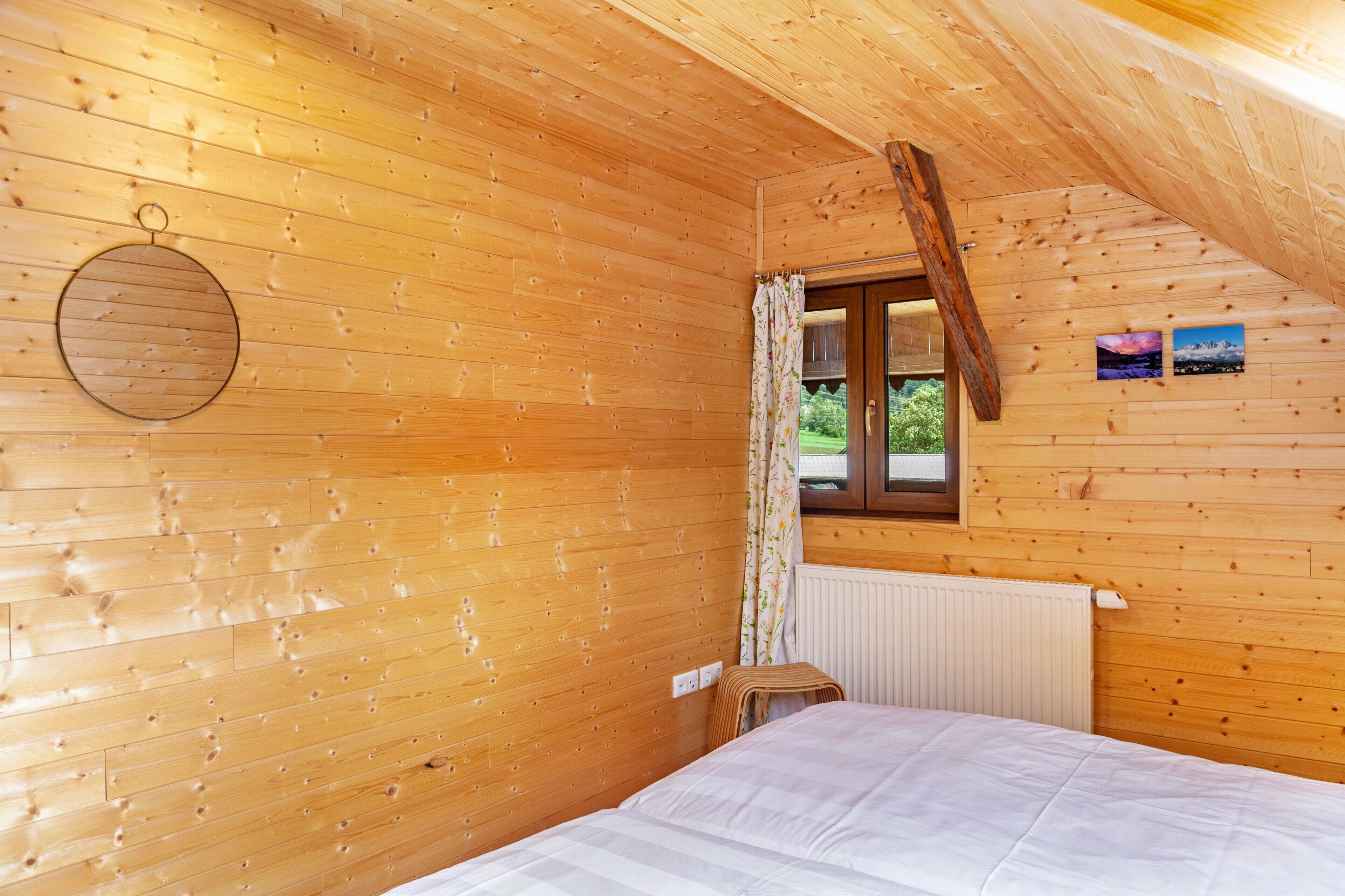 Una camera di una baita in legno con un letto, una finestra con tende, un termosifone e un quadro appeso al muro.