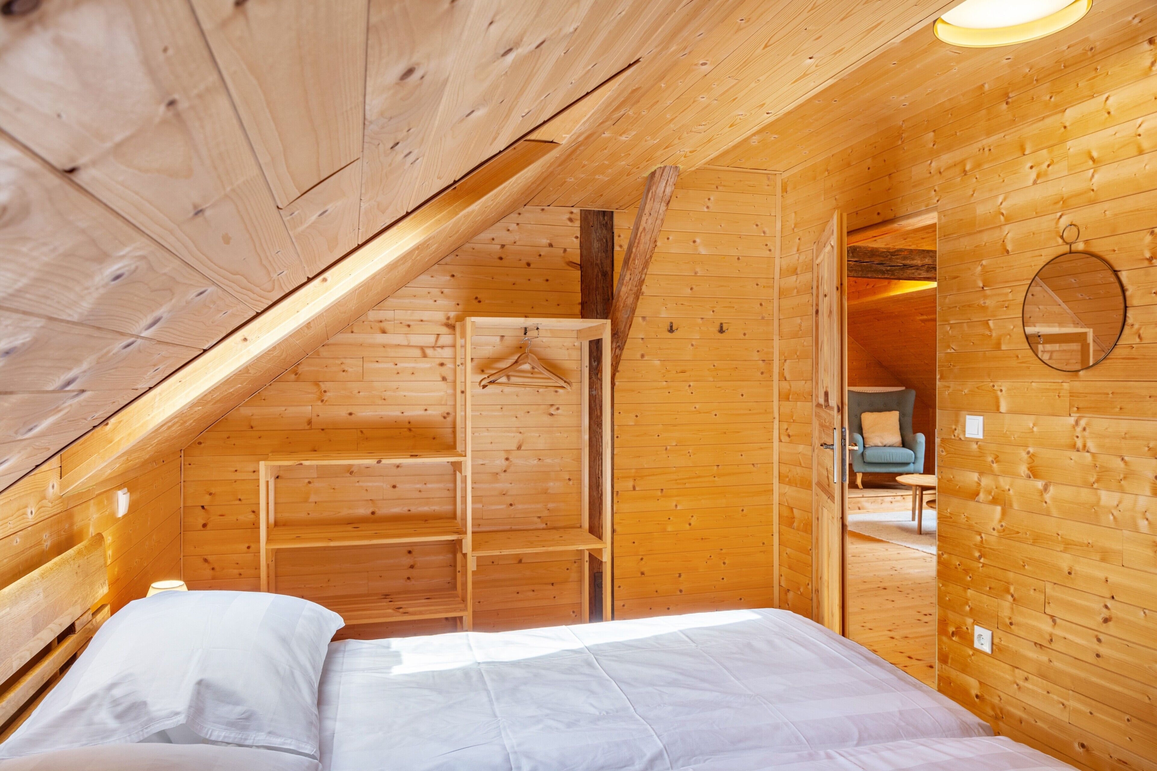 A wooden cabin bedroom with a sloped ceiling, a bed, a hanging coat rack, and a small seating area with a chair and table.