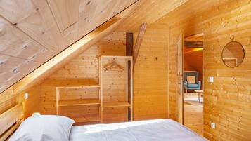 A wooden cabin bedroom with a sloped ceiling, a bed, a hanging coat rack, and a small seating area with a chair and table.