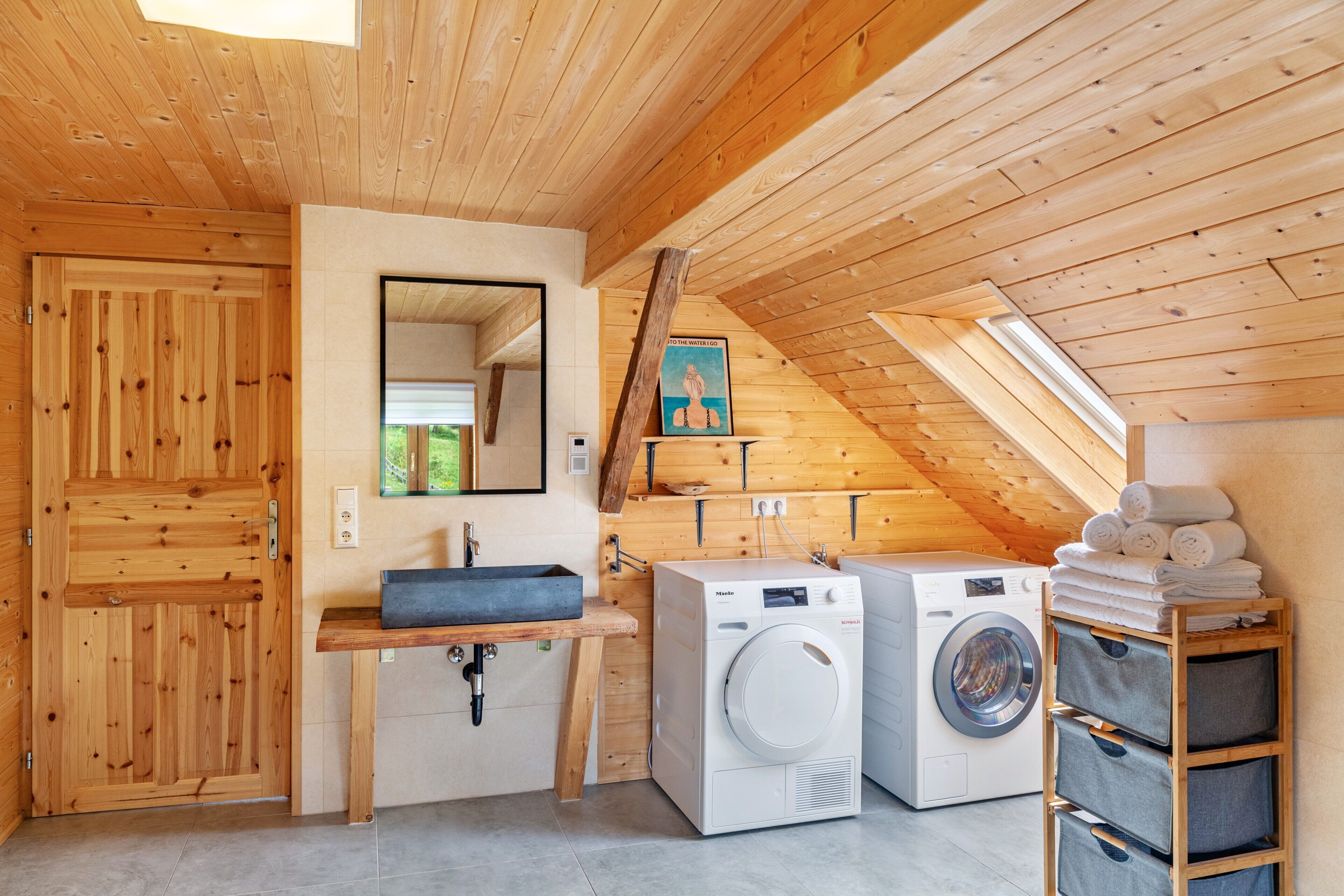 A compact, wooden-floored laundry room with a washing machine, dryer, and a shelf with folded towels.