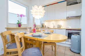 A compact kitchen with modern appliances, a dining area with wooden furniture, and a large window with blinds.