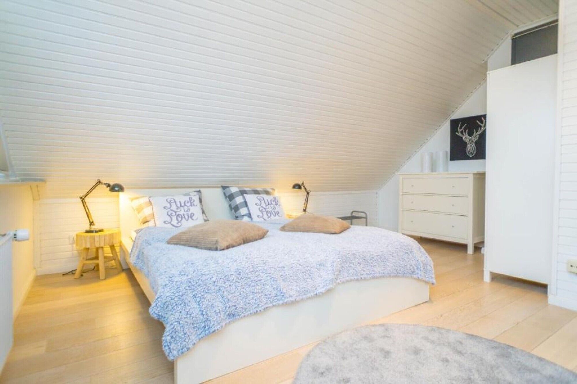 A bedroom with a sloped ceiling, a bed with a white and blue patterned bedspread, two bedside lamps, a dresser, and a wall-mounted deer head decoration.