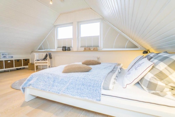 A bedroom with a sloped ceiling, a bed with a blue blanket, two windows with blinds, and a radiator.