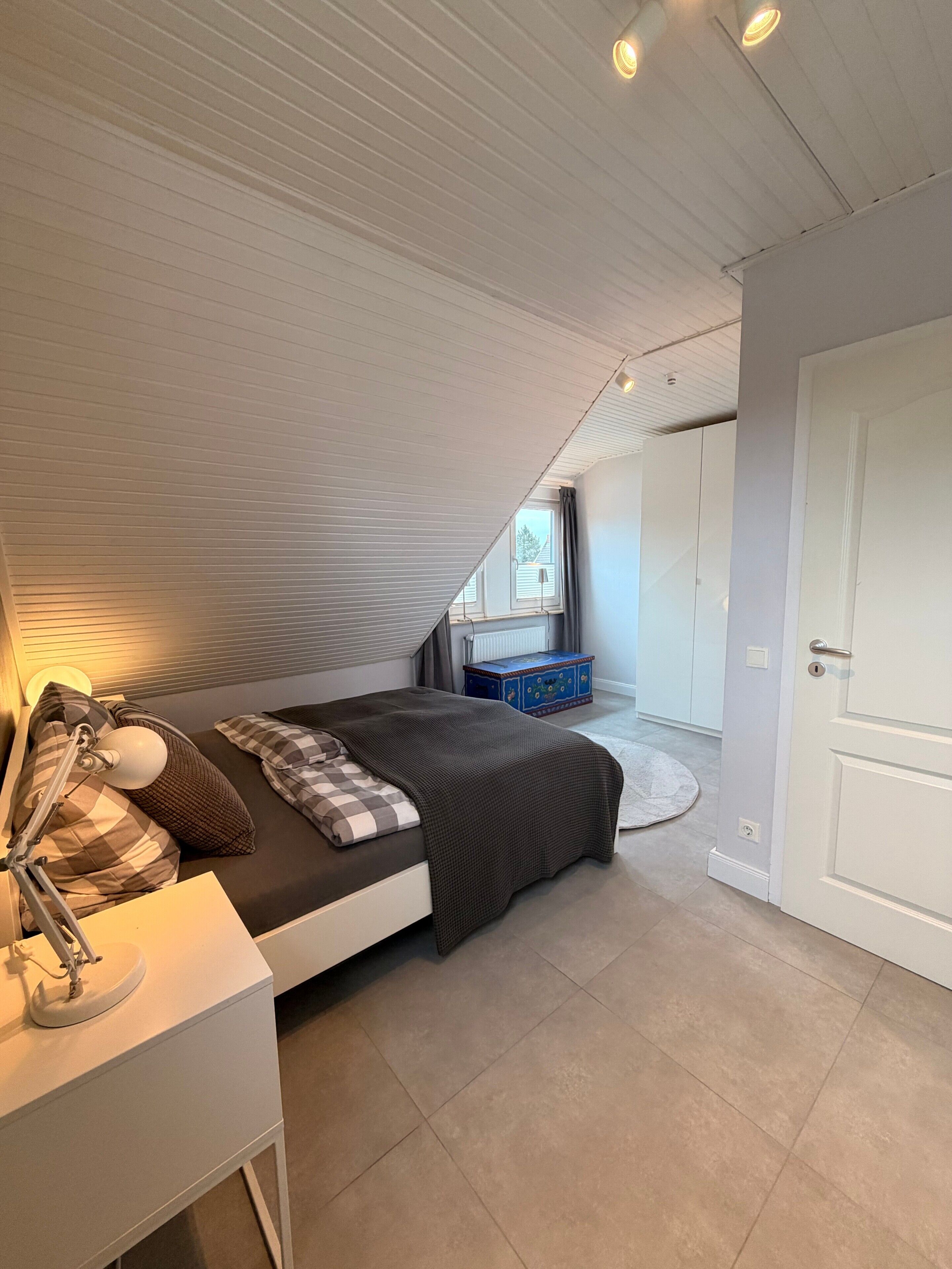 A bedroom with a sloped ceiling, a bed with a checkered blanket, a bedside table with a lamp, a dresser, and a window with curtains.
