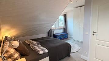 A bedroom with a sloped ceiling, a bed with a checkered blanket, a bedside table with a lamp, a dresser, and a window with curtains.