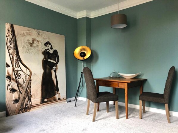 A dining area with a table, chairs, a lamp, and a large painting on the wall.