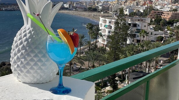 A balcony with a white pineapple sculpture, a blue cocktail glass with fruit garnish, and a coastal cityscape in the background. - Ocean View 2 (Los Cristianos)