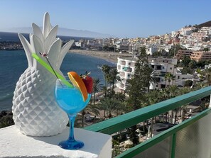 A balcony with a white pineapple sculpture, a blue cocktail glass with fruit garnish, and a coastal cityscape in the background. - Ocean View 2 (Los Cristianos)