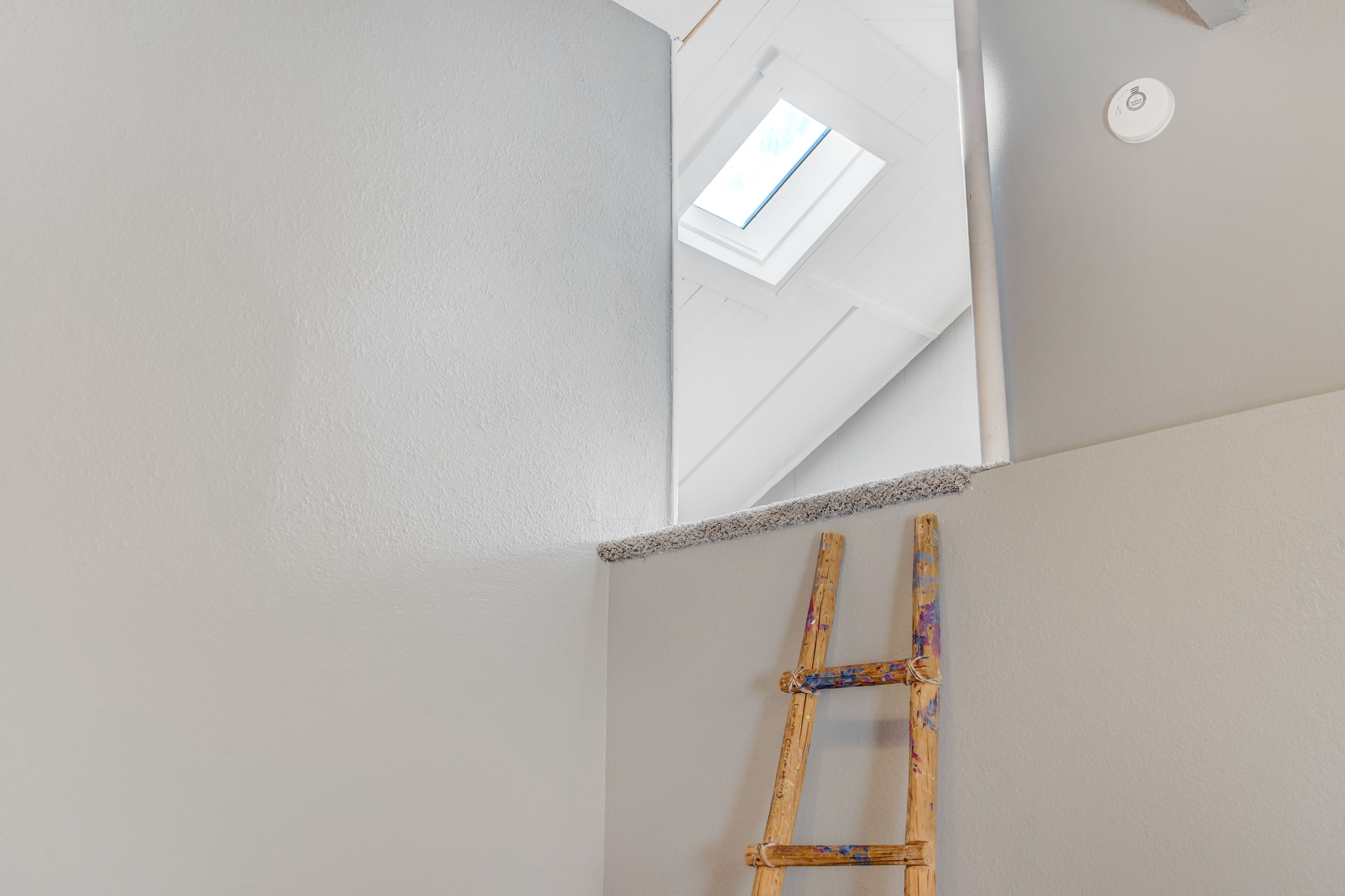 A corner of a room with a white wall, a skylight, and a wooden ladder leaning against the wall.